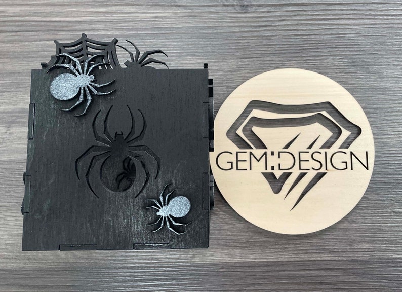 May include: Black wooden box with a spider web design and three silver spiders. The box is next to a round wooden sign with a diamond shape and the text "GEM:DESIGN".