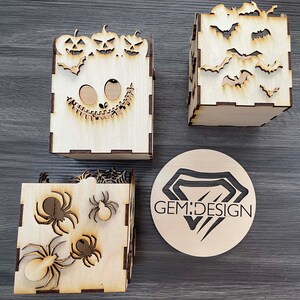 May include: Three wooden boxes with Halloween themes. The first box features a jack-o-lantern face with three pumpkins on top. The second box has a spider web and spiders. The third box has bats. The boxes are all made of wood and have a natural finish. The boxes are all approximately 10 cm tall and 10 cm wide. GEM:DESIGN is printed on a round wooden disc.