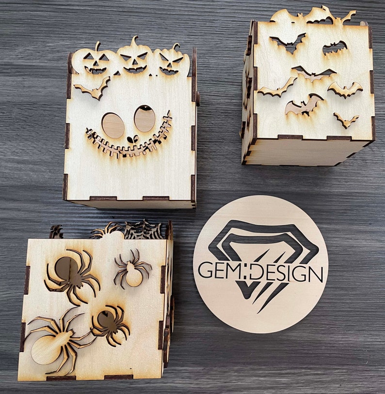May include: Three wooden boxes with Halloween themes. The first box features a jack-o-lantern face with three pumpkins on top. The second box has a spider web and spiders. The third box has bats. The boxes are all made of wood and have a natural finish. The boxes are all approximately 10 cm tall and 10 cm wide. GEM:DESIGN is printed on a round wooden disc.