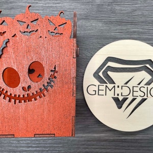 May include: A wooden box shaped like a jack-o'-lantern with three smaller pumpkins on top. The box has a stitched mouth and eyes. The box is painted orange and has a black bat on the front. A round wooden sign with the text "GEM:DESIGN" is in the background.