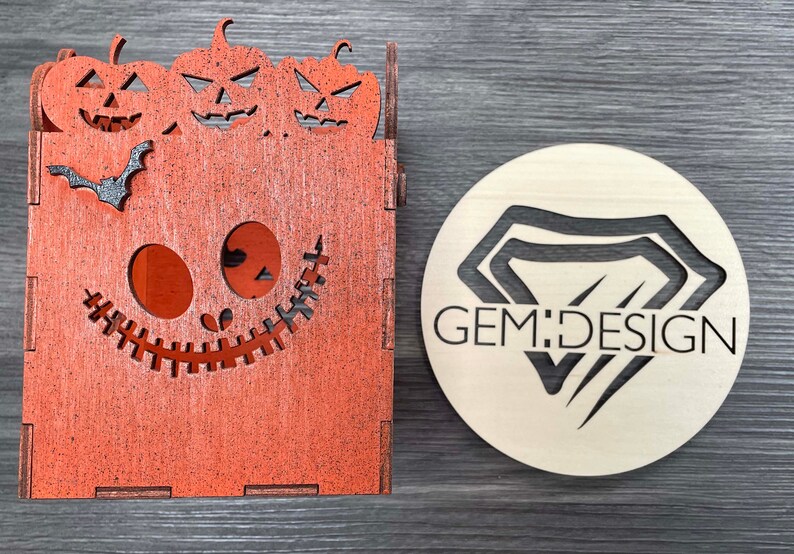May include: A wooden box shaped like a jack-o'-lantern with three smaller pumpkins on top. The box has a stitched mouth and eyes. The box is painted orange and has a black bat on the front. A round wooden sign with the text "GEM:DESIGN" is in the background.