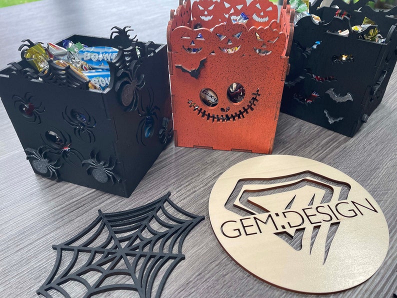 May include: Three Halloween-themed wooden boxes with sweets inside. The boxes are decorated with spiders, bats, and jack-o'-lanterns. The boxes are sitting on a wooden surface. The boxes are labelled "GEM:DESIGN".