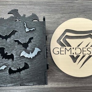 May include: A black wooden box with a cutout design of bats. The box has a distressed finish and is decorated with silver glitter. The box is next to a wooden circle with a cutout design of a diamond and the text "GEM:DESIGN".