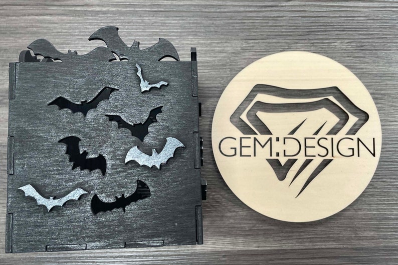 May include: A black wooden box with a cutout design of bats. The box has a distressed finish and is decorated with silver glitter. The box is next to a wooden circle with a cutout design of a diamond and the text "GEM:DESIGN".