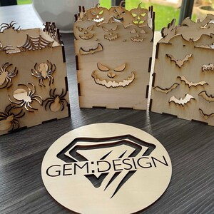 May include: Three wooden boxes with Halloween themes. The first box has a spider web design with spiders. The second box has a jack-o-lantern face. The third box has a bat design. The boxes are all made of natural wood and have a rustic look. GEM:DESIGN is printed on a round wooden disc.