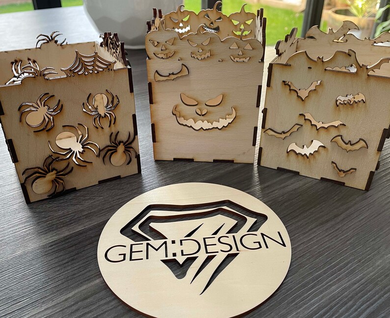 May include: Three wooden boxes with Halloween themes. The first box has a spider web design with spiders. The second box has a jack-o-lantern face. The third box has a bat design. The boxes are all made of natural wood and have a rustic look. GEM:DESIGN is printed on a round wooden disc.