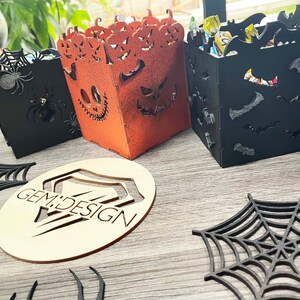 May include: Three Halloween-themed wooden boxes with different designs. The first box is black with a spider web and spiders. The second box is orange with a jack-o'-lantern face. The third box is black with bats. The boxes are on a wooden surface with a wooden cutout of a spider web and the words "GEM DESIGN".