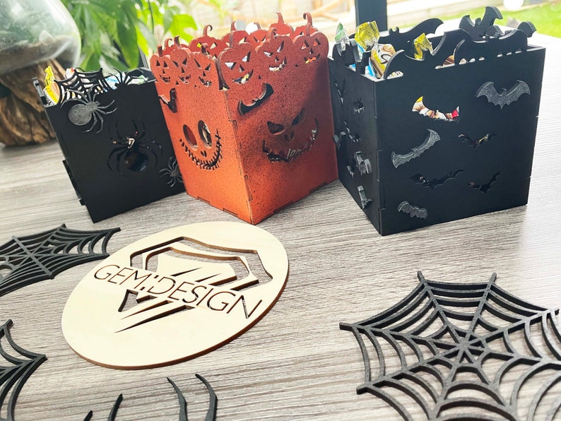 May include: Three Halloween-themed wooden boxes with different designs. The first box is black with a spider web and spiders. The second box is orange with a jack-o'-lantern face. The third box is black with bats. The boxes are on a wooden surface with a wooden cutout of a spider web and the words "GEM DESIGN".