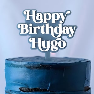 May include: A two-tiered blue cake with a white cake topper that says "Happy Birthday Hugo" on a white cake stand.