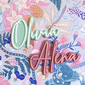 May include: Two names, "Olivia" and "Aiena", are written in a cursive font on a light blue background with a floral pattern. The names are cut out of wood and painted in different colours. "Olivia" is painted in a light green colour and "Aiena" is painted in a light pink colour.