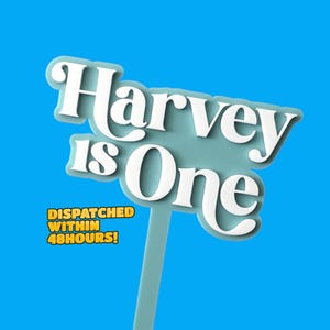 May include: White cake topper with the text "Harvey is One" in a blue and white colour scheme. The topper is on a blue background.