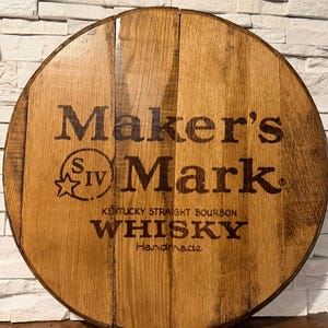 May include: Round wooden sign made from a barrel head. The sign features the words "Maker's Mark" in large, dark letters, along with "Kentucky Straight Bourbon Whisky" and "Handmade". The wood has a natural, aged appearance.