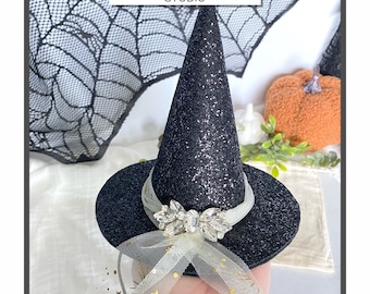 Puede incluir: Un sombrero de bruja negro con purpurina que presenta una banda de cinta de tul blanco adornada con un adorno de cristal transparente y pequeños detalles de estrellas doradas. El texto Foxie Boutique STUDIO aparece arriba.