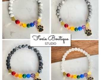May include: Four bracelets with rainbow charm and paw print charm. The bracelets are made with white, gray, and black stones. The charms are gold with black accents. The text "Foxie Boutique STUDIO" is visible in the image.