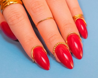 May include: Close-up of a hand with red nail polish and gold rings. The nails are long and pointed, and the rings are simple and elegant.