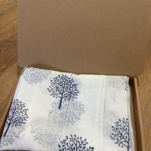 May include: A white fabric square featuring a repeating pattern of dark blue trees with detailed branches and leaves, neatly folded and placed inside an open brown cardboard box with a hinged lid, presenting a clean and organised appearance.