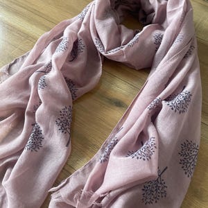 May include: A dusky pink scarf featuring a repeating tree design in a darker hue. The scarf is draped and folded, with fringed edges. The fabric appears lightweight and soft, suitable for various styles.