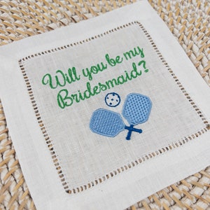 Peut inclure: Serviette en lin blanc avec une broderie verte qui dit "Veux-tu &ecirc;tre ma demoiselle d'honneur ?" et deux raquettes de paddleball brod&eacute;es en bleu.