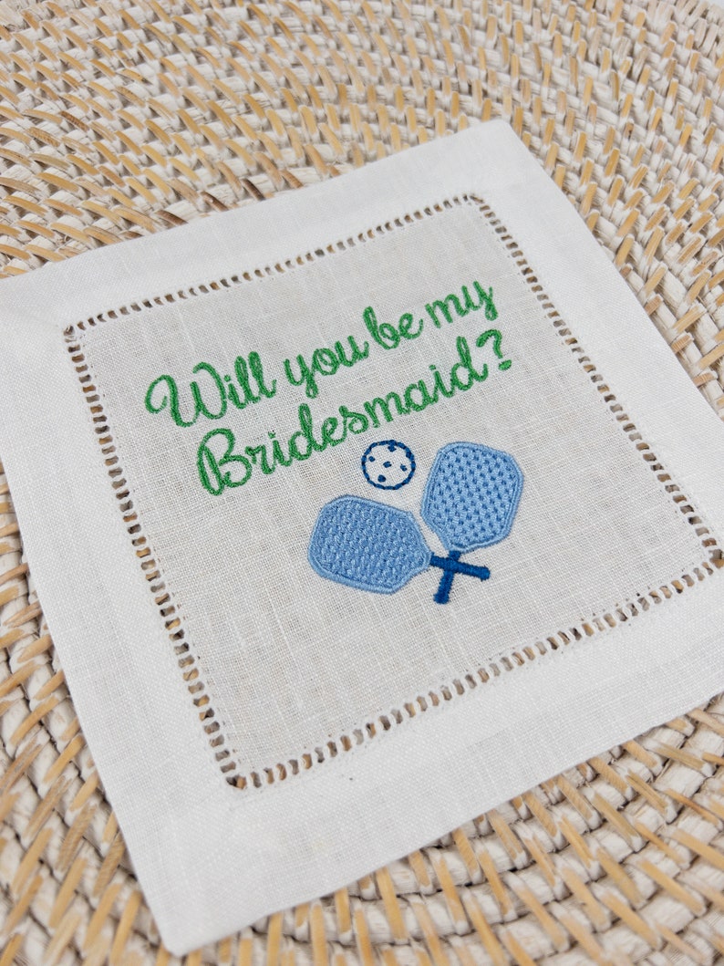 Peut inclure: Serviette en lin blanc avec une broderie verte qui dit "Veux-tu &ecirc;tre ma demoiselle d'honneur ?" et deux raquettes de paddleball brod&eacute;es en bleu.