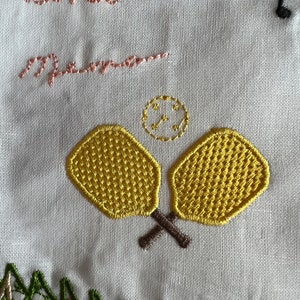 Peut inclure: Broderie sur tissu blanc avec les mots "Mama" et "mamma" en fil rose, deux raquettes de paddle jaunes avec des manches brunes et un petit cercle avec une croix &agrave; l'int&eacute;rieur en fil dor&eacute;.