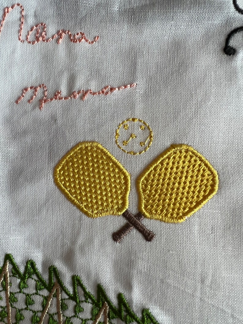 Peut inclure: Broderie sur tissu blanc avec les mots "Mama" et "mamma" en fil rose, deux raquettes de paddle jaunes avec des manches brunes et un petit cercle avec une croix &agrave; l'int&eacute;rieur en fil dor&eacute;.