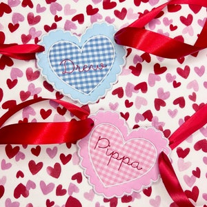 May include: Two heart-shaped fabric tags with gingham patterns and scalloped edges. The blue tag says "Drew" and the pink tag says "Pippa". Both tags are tied with red ribbon on a white background with pink and red hearts.