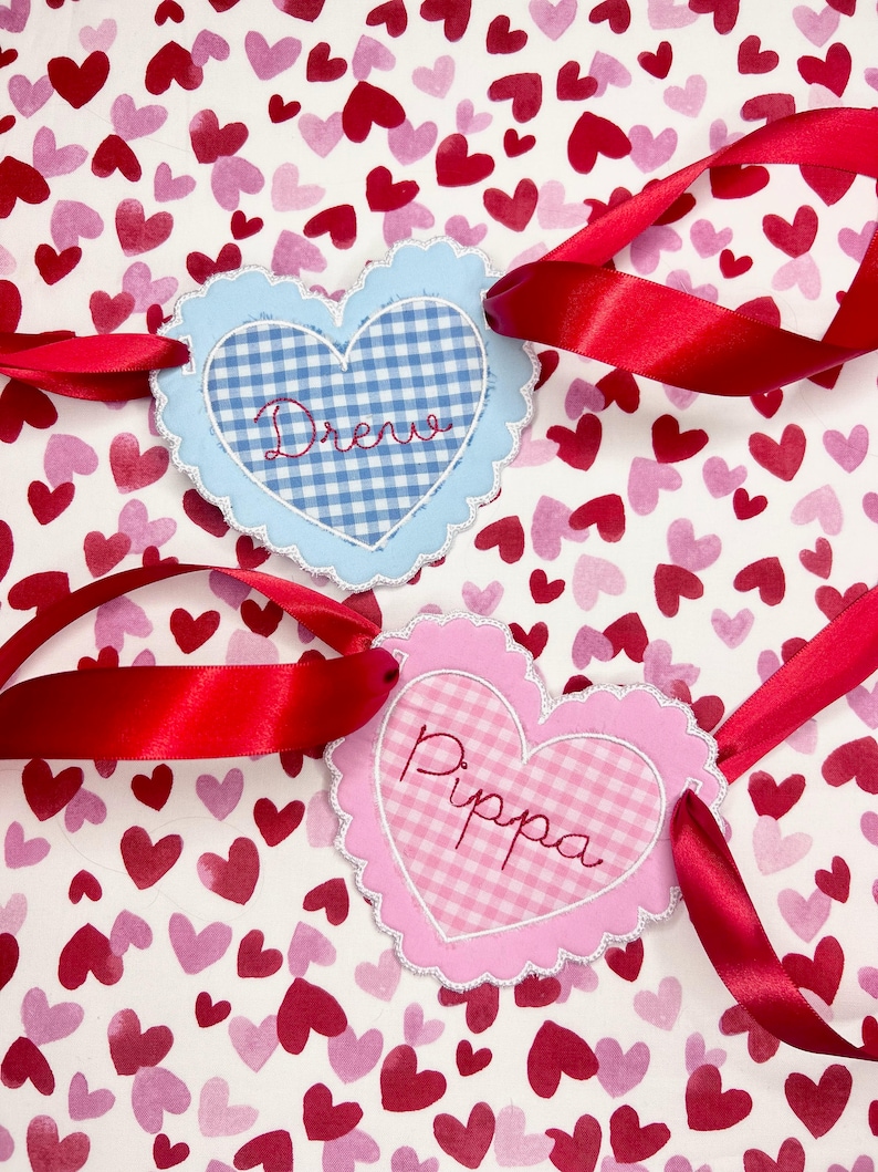 May include: Two heart-shaped fabric tags with gingham patterns and scalloped edges. The blue tag says "Drew" and the pink tag says "Pippa". Both tags are tied with red ribbon on a white background with pink and red hearts.