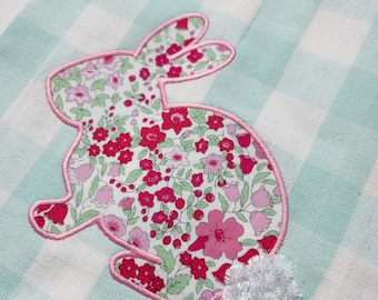 May include: A teal and white gingham kitchen towel with a pink floral bunny applique. The bunny has a white fluffy tail.