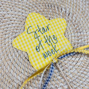 May include: A yellow and white gingham star-shaped award with the text "Star of the Week" in blue embroidery. The star is attached to a wooden stick with blue and white gingham ribbon and a gold ribbon.