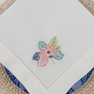 Peut inclure: Serviette en lin blanc avec un motif de fleur brodé coloré. La fleur est rose, jaune, verte et bleue.