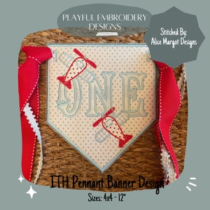 May include: A blue and white pennant banner with the word "ONE" embroidered in a script font. Two red and white airplanes are embroidered on either side of the number. The banner is 4 inches by 4 inches and 12 inches long. The banner is on a woven wicker background.