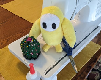 May include: A yellow plush toy shaped like a character from the game Among Us, a green cactus, a pair of scissors, and a bottle of 3-in-1 advanced craft glue on a white sewing machine table.