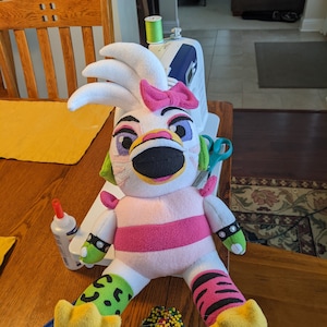 以下が含まれることがあります： 黄色いクチバシ、緑色の目、ピンクのリボンが付いたピンクと白のぬいぐるみ。おもちゃは、糸巻き、針山、ミシンなど、裁縫道具が置かれた木製のテーブルの上に置かれています。