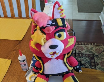 May include: A red and yellow plush toy with a black and white striped jacket, a pink head, and a yellow star on its eye. The toy is sitting on a wooden table with sewing supplies.