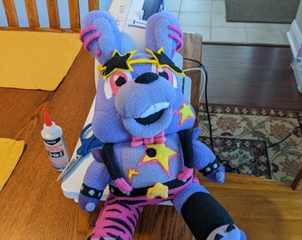 May include: A purple and pink plush toy with a yellow star on its chest and a black and white striped pattern on its legs. The toy is sitting on a wooden table with a sewing machine in the background.