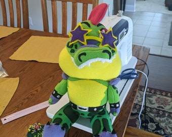 May include: A green and yellow plush crocodile wearing sunglasses and a black studded belt. The crocodile is sitting on a white surface with a sewing machine and scissors in the background.