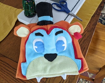 May include: A blue and orange plush toy hat with a black top hat, white eyes, and a black nose. The hat is designed to look like a cartoon animal.