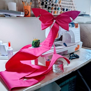 Könnte beinhalten: Eine Nähmaschine mit einer großen rosa Schleife, die darüber drapiert ist. Die Maschine hat einen Halloween-Kürbis-Aufkleber. Ein kleines Nadelkissen und anderes Nähzubehör befinden sich auf dem Tisch. Eine Schreibtischlampe befindet sich links.