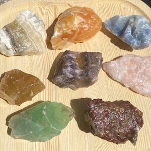 May include: A collection of eight rough, natural crystals in various colors, including green, orange, pink, purple, brown, and blue. The crystals are arranged on a wooden surface.
