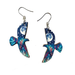 May include: A pair of silver dangle earrings featuring a blue and green bird with a full moon and stars in the background.
