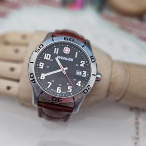 Könnte beinhalten: Eine Wenger-Uhr mit schwarzem Zifferblatt, silberner Lünette und braunem Lederarmband. Die Uhr hat weiße Ziffern, ein Datumsfenster und einen roten Sekundenzeiger. Swiss Made.