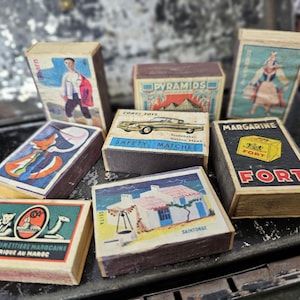 May include: A collection of vintage matchboxes with colorful illustrations. The boxes feature images of people, cars, and buildings. Some boxes have text, including "SAFETY MATCHES" and "FORT MARGARINE".