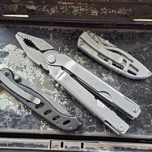 Peut inclure: Un outil multifonction Gerber argenté avec pinces intégrées et une règle graduée en centimètres, accompagné de deux couteaux pliants avec manches métalliques et clips de poche, le tout avec une finition mate et des vis apparentes.