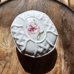 May include: White ceramic trinket box with a woven basket design. The lid features an oval-shaped floral design with a pink and red lily. The box is set on a wooden tray.