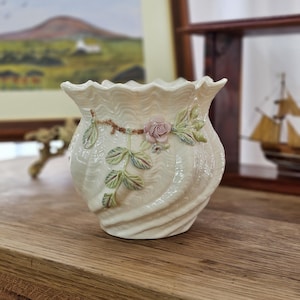 May include: Cream-colored ceramic vase with a ruffled rim and a swirling textured design. The vase is adorned with a pink rose and green leaves. A decorative piece for home decor.