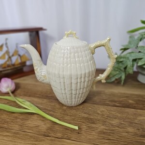 Puede incluir: Tetera de cerámica color crema con un cuerpo texturizado y acanalado y un asa en forma de rama. La tetera tiene una tapa a juego y un pico curvo. Un tulipán decorativo y una maqueta de barco están en el fondo.