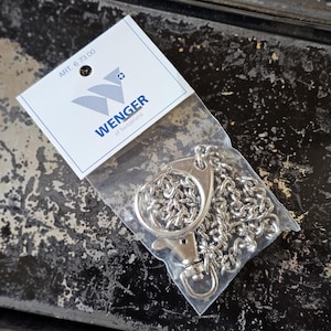 Puede incluir: Una cadena de metal plateado con un cierre giratorio, empaquetada en una bolsa de plástico transparente con una tarjeta de cabecera blanca. La tarjeta muestra el logo azul de Wenger of Switzerland y el texto ART. 6 73 00 en tinta negra.