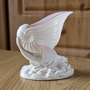 May include: A white ceramic sculpture of a fish with a large, fan-shaped fin and a nautilus shell. The sculpture has a pink and gold accent on the fin and shell. The fish rests on a base of stylized waves.