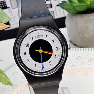 May include: A black Swatch Quartz watch with a white dial featuring black numerals and a black center. The watch has a black strap and colorful hands: blue, yellow, and red. The watch face reads "Swatch Quartz".