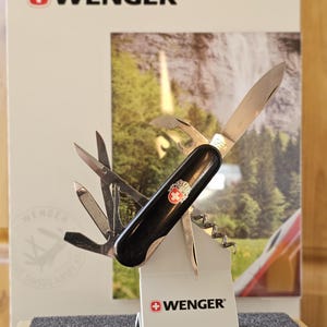 Pode incluir: Um canivete multifuncional Wenger preto com várias ferramentas de aço inoxidável abertas, incluindo uma lâmina principal, tesoura e saca-rolhas. O cabo apresenta um emblema de cruz suíça vermelha e branca. A marca WENGER está impressa no suporte.